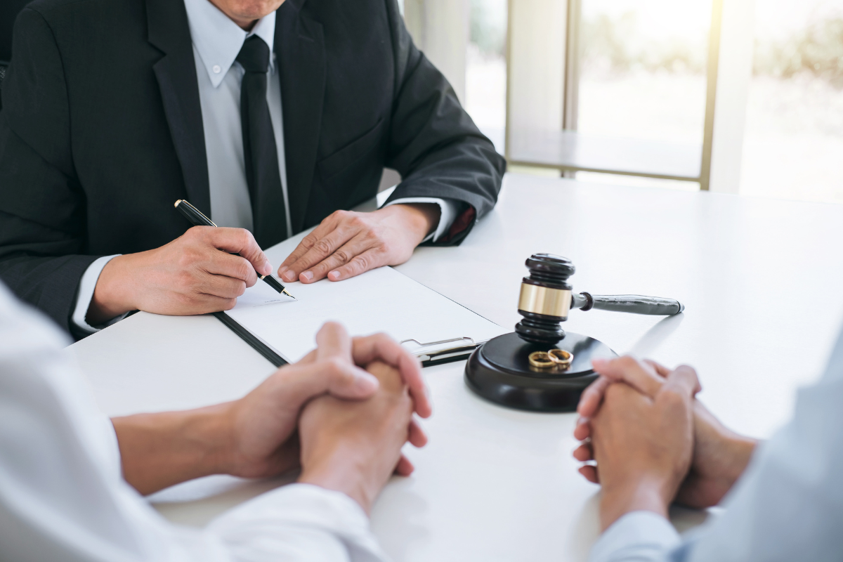 Two people sit across from a third person signing papers, there is a gavel in front of the third person, signifying that he is an attorney, the two people sitting across the third are finalizing their divorce